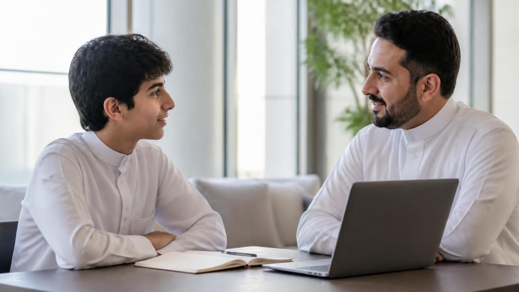 A Saudi teenage boy wearing a white thobe sits with an adult male mentor in a bright, modern space, with a notebook and laptop on the table.